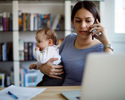mom holding baby while on cell phone working on computer