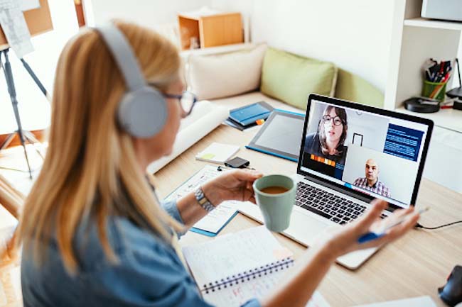 A virtual interview being held. A woman is holding a coffee cup at home speaking to two people on her laptop.