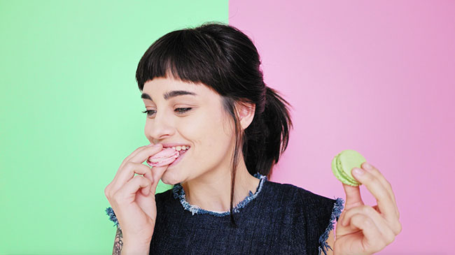 Woman in the center of a green and pink wall eating a pink macaroon while holding a green macaroon.