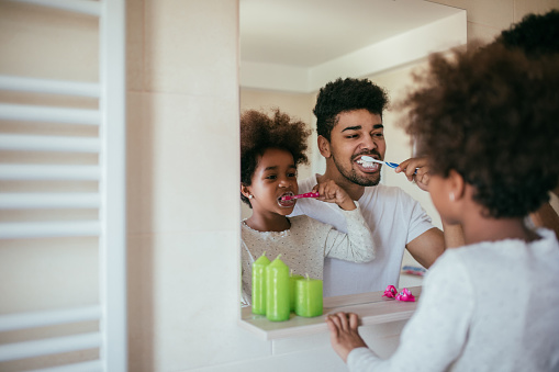 Portrait of black dad and daughter brushing teeth and looking in the mirror in the bathroom