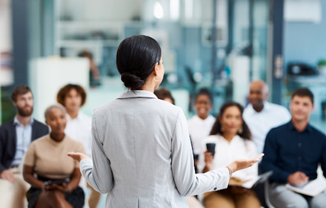 Company lead holding an employee training session to improve customer satisfaction scores. A woman in a gray suit standing in front of an audience.