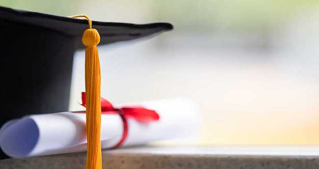 A close-up image of a black graduation cap with golden tassle and a white rolled-up diploma with a red ribbon.