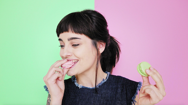 Curion: Woman eating pink and green macaroons with a green and pink background. 